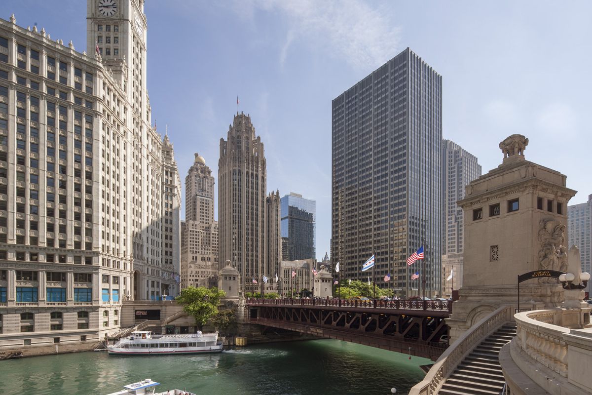 Optima Lakeview® Apartments in Chicago, IL A boat passes under a bridge on the Chicago River, embracing blue space amidst tall city buildings on a sunny day.