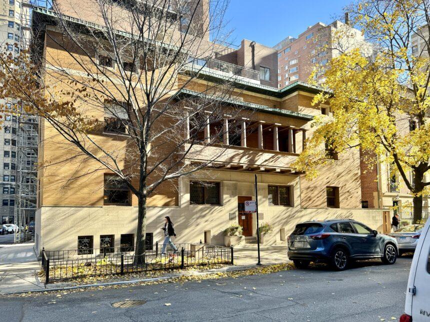 Optima Lakeview® Apartments in Chicago, IL A person walks past a tan, modern-style building on a sunny street, discovering Chicago among autumn trees and parked cars.