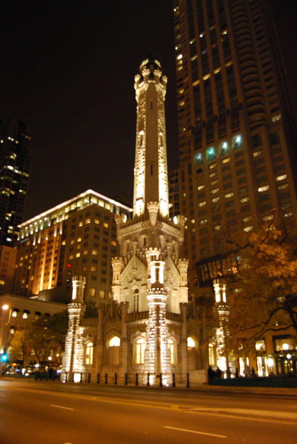 Optima Lakeview® Apartments in Chicago, IL A tall, illuminated historic stone tower stands among modern buildings at night in an urban setting.