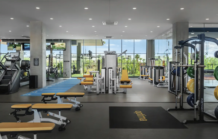 Optima Lakeview® Apartments in Chicago, IL Modern gym with yellow benches, Optima weight machines, cardio equipment, and large windows overlooking seasonal greenery.