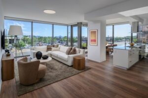 Optima Lakeview® Apartments in Chicago, IL Modern living room with radiant sunlight, large windows, neutral furniture, hardwood floors, and views of trees and buildings outside.