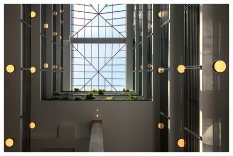 Optima Lakeview® Apartments in Chicago, IL View looking up at a skylight with round wall lights and green plants on a ledge, bright natural light above—pure Optima Way living.