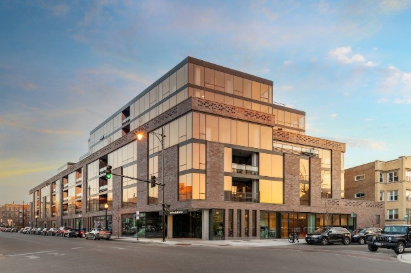 Optima Lakeview® Apartments in Chicago, IL Modern multi-story building with large glass windows on a city corner at sunset, cars parked along the street.