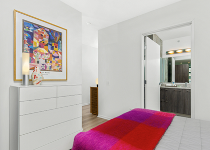 Optima Lakeview® Apartments in Chicago, IL Bright bedroom with colorful blanket, white dresser, abstract art, and view into a modern bathroom.