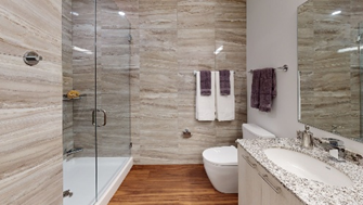 Optima Lakeview® Apartments in Chicago, IL Modern bathroom with glass shower, granite countertop, wood floor, and purple and white towels on the wall.