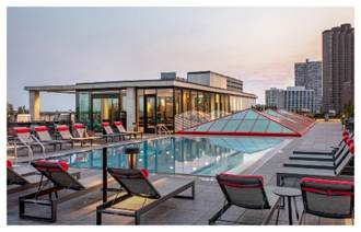 Optima Lakeview® Apartments in Chicago, IL Rooftop pool with lounge chairs, glass skylights, and city buildings in the background at sunset.