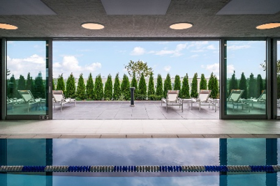 Optima Lakeview® Apartments in Chicago, IL Indoor pool with blue lane dividers at Optima, facing lounge chairs and greenery beyond large glass doors to outdoor space.