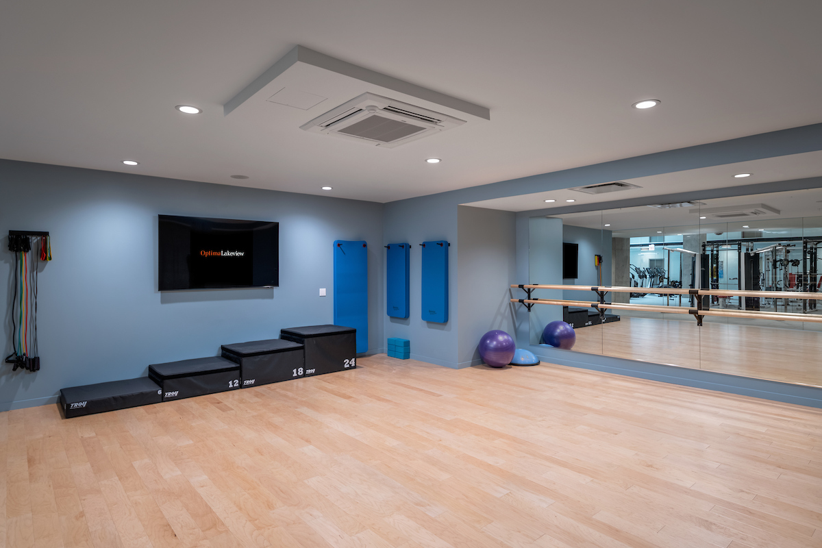 Optima Lakeview® Apartments in Chicago, IL Modern fitness studio at Optima Lakeview with wooden floors, Pilates gear, wall mirror, mounted TV, and blue yoga mats on a gray wall.
