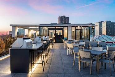 Optima Lakeview® Apartments in Chicago, IL Rooftop restaurant at sunset with tables, chairs, bar seating, and city buildings in the background.