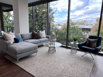 Optima Lakeview® Apartments in Chicago, IL Modern living room with a gray sofa, armchair, glass tables, rug, and large windows overlooking trees and sky.